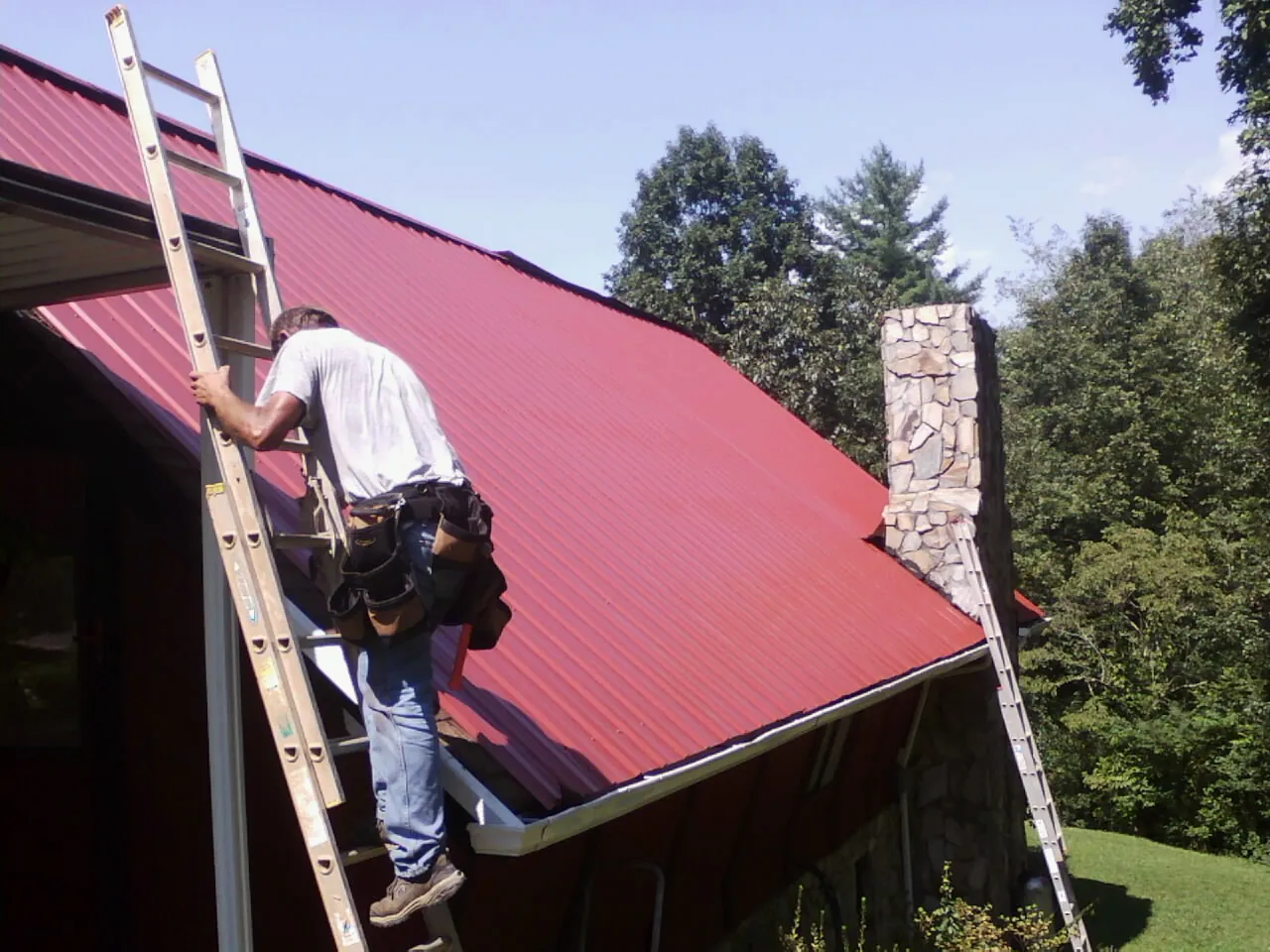 Roofer on a ladder beside a red metal roof and stone chimney — Soffit & Fascia Repair by Roofing Brothers in Tenafly